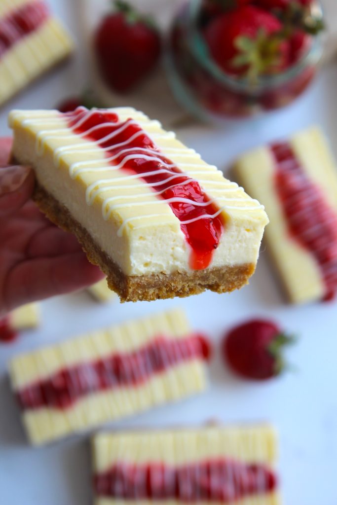 A close-up of a hand holding a cheesecake bar with a graham cracker crust, topped with white chocolate drizzle and a streak of strawberry sauce. Other similarly decorated cheesecake bars and fresh strawberries are visible in the background.