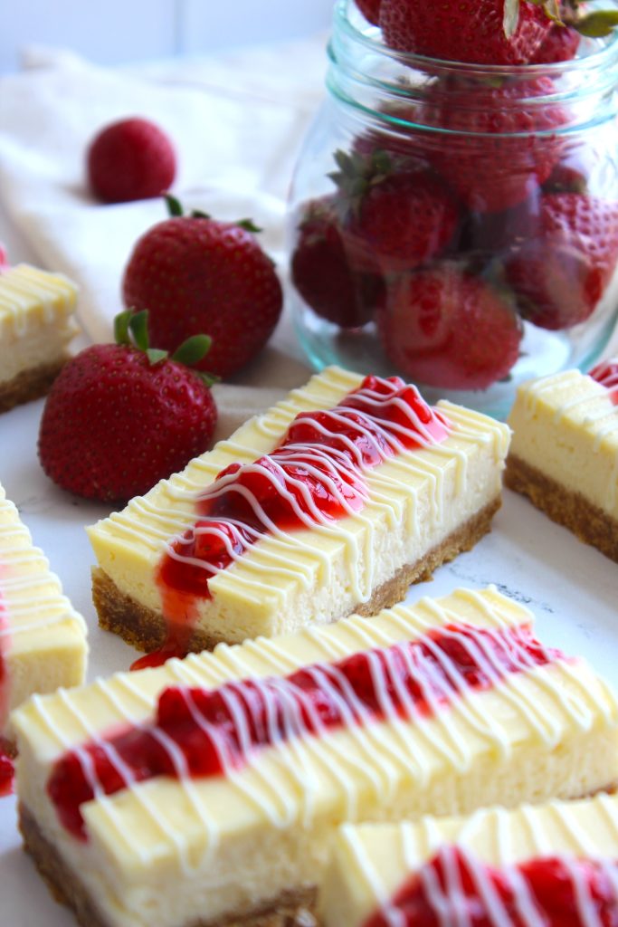A close-up of rectangular cheesecake bars topped with a red strawberry sauce and white drizzle, arranged on a white surface. Fresh strawberries and a glass jar filled with more strawberries are in the background.