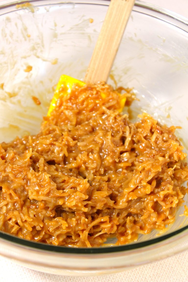 A glass bowl containing a sticky mixture of ingredients with a wooden spatula, likely in the process of preparing a dessert or confectionery like rice krispie treats or caramelized treats.