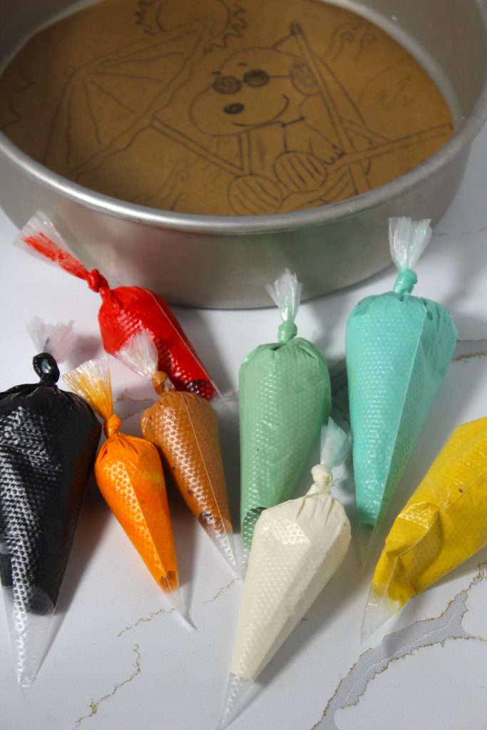 Assorted colorful piping bags filled with sponge cake batter, arranged on a table. The bags range in colors like black, red, green, orange, white, and yellow.