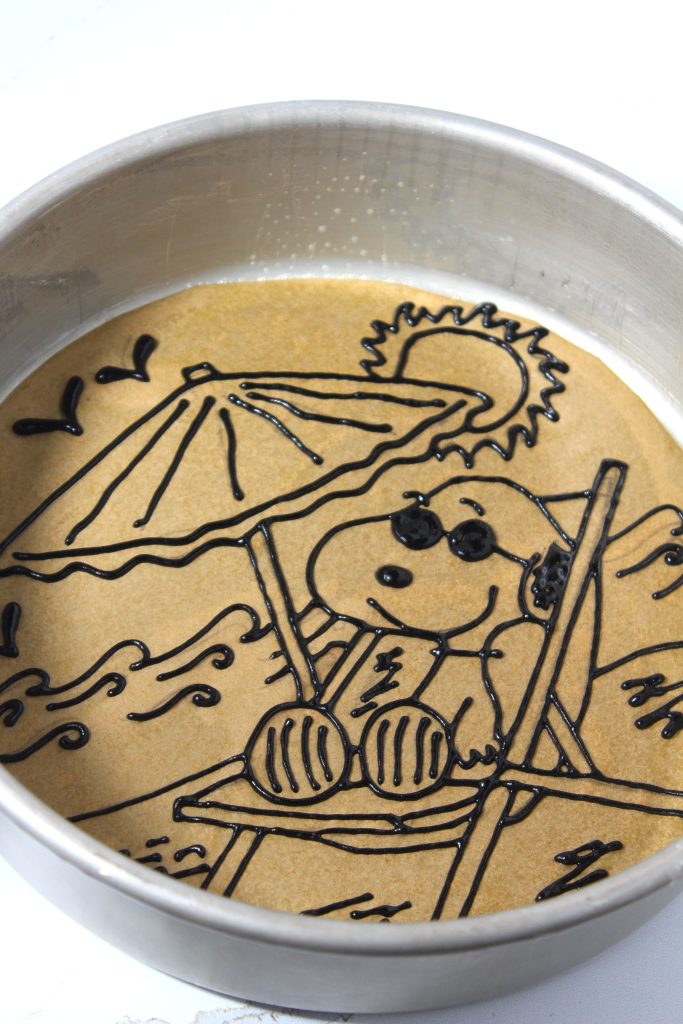 Black sponge cake batter pipied ver an image of snoopy on parchment paper in a circular cake pan.