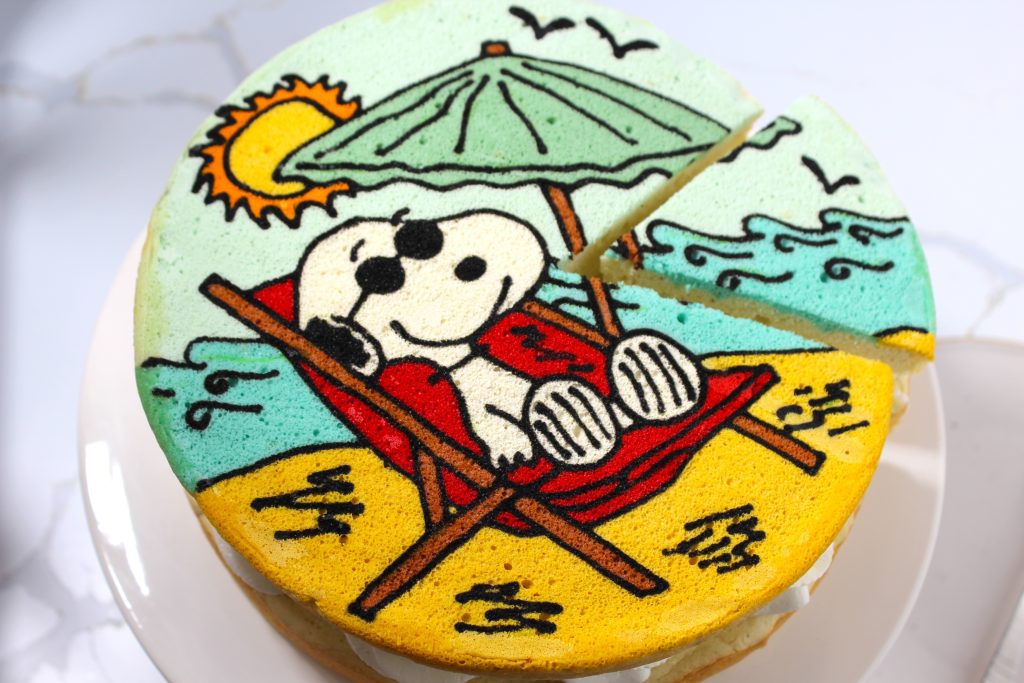 A decorated cake featuring the character snoopy from the peanuts comic strip, lounging in a red chair under an umbrella on a beach, with sunny sky and ocean waves depicted in icing.