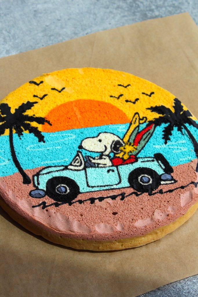 A round cake decorated with an illustration of Snoopy and Woodstock driving in a blue car. The background features a sunset with orange and yellow hues, palm trees, birds, and an ocean view. The cake is placed on a piece of parchment paper.