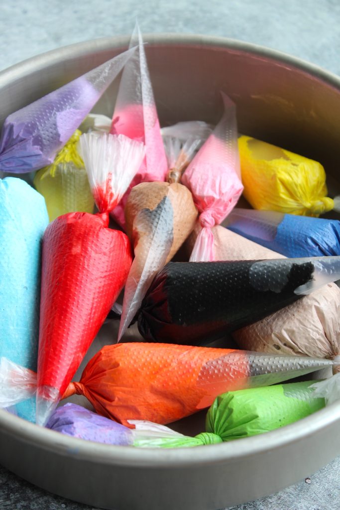 A round metal container holds a variety of filled piping bags tipped with different vibrant colors, including red, blue, black, orange, purple, pink, yellow, and brown. The bags are arranged in a visually appealing manner, suggesting they are ready for use in cake decorating.