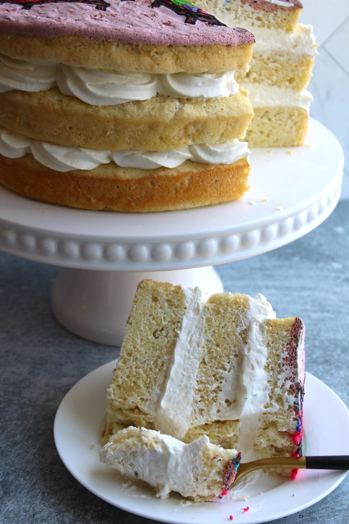 A layered vanilla cake with white frosting is displayed on a white cake stand. A slice has been served on a white plate with a fork, showing the cake's fluffy texture and thick frosting layers. The top of the cake has a hint of purple decoration.
