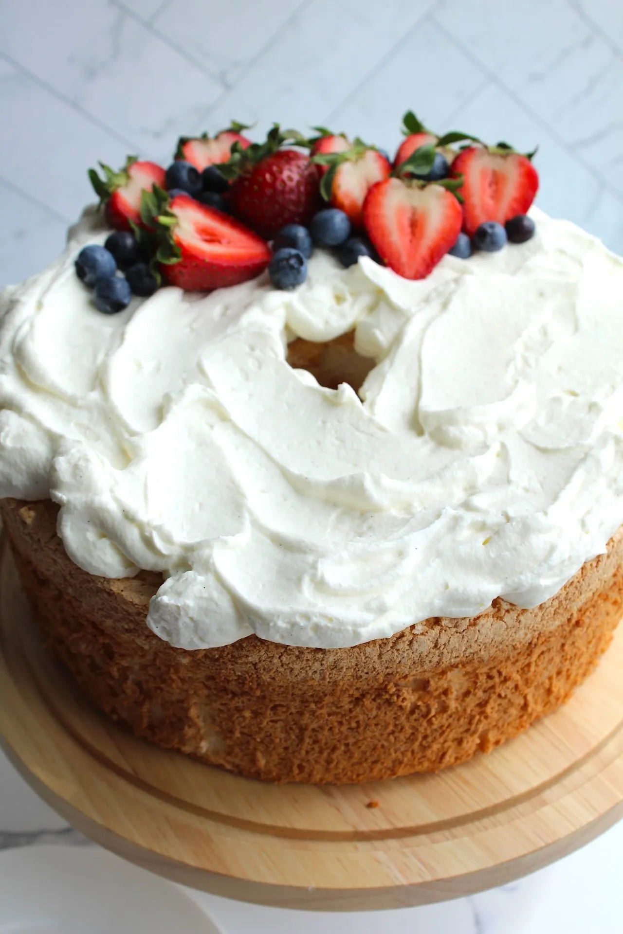 A round sponge cake with white frosting is topped with a mix of fresh fruits, including halved strawberries and blueberries. The cake is centered on a wooden cake stand placed on a marble surface.