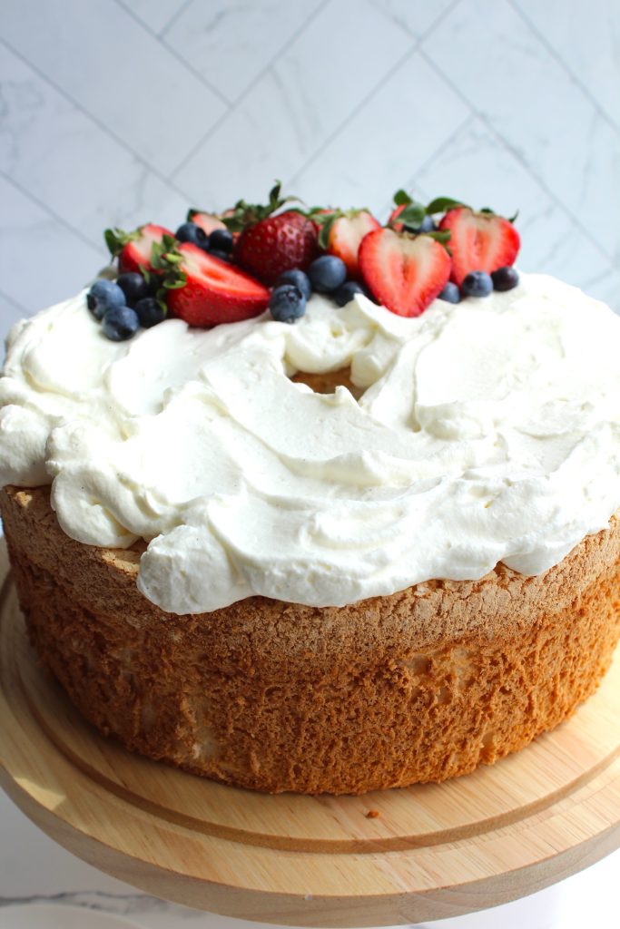 A round angel food cake topped with a generous layer of white whipped cream and garnished with sliced strawberries, whole blueberries, and small green leaves. The cake is placed on a light wooden cake stand against a white tiled background.