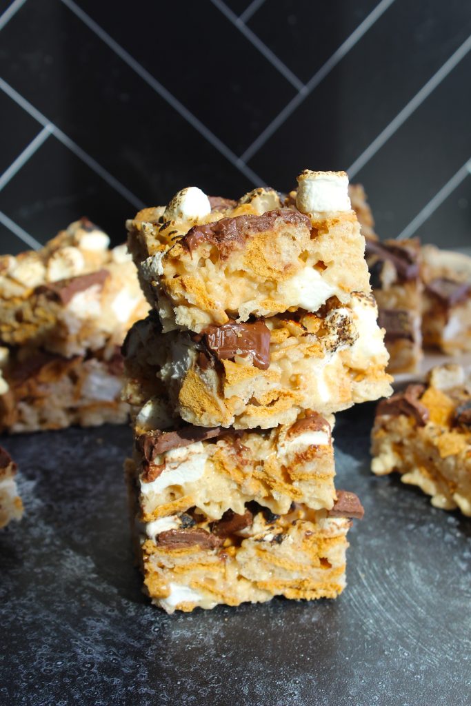 A stack of three s'mores bars sits on a dark surface with more bars in the background. The bars are made with pieces of chocolate, marshmallows, and graham crackers, creating a layered and textured appearance. A black and white tiled wall is visible behind them.