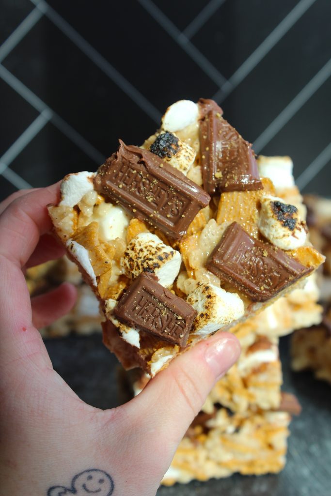 A hand holding a homemade s'mores bar topped with toasted marshmallows and chocolate chunks. The bar also includes graham crackers and a gooey golden brown base. In the background, more s'mores bars are stacked on a black-tiled surface.