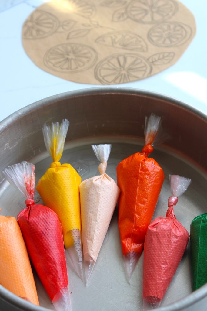 A round metal tray holds eight piping bags filled with brightly colored icing, including red, orange, yellow, and green. In the background, there is a large brown piece of rolled-out dough with designs of circular citrus slices printed on it.