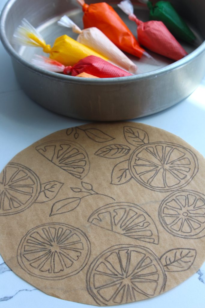 A metal baking pan containing colorful wrapped items sits on a white marble surface. In front of the pan is a round piece of brown parchment paper with black ink drawings of sliced citrus fruits and leaves.