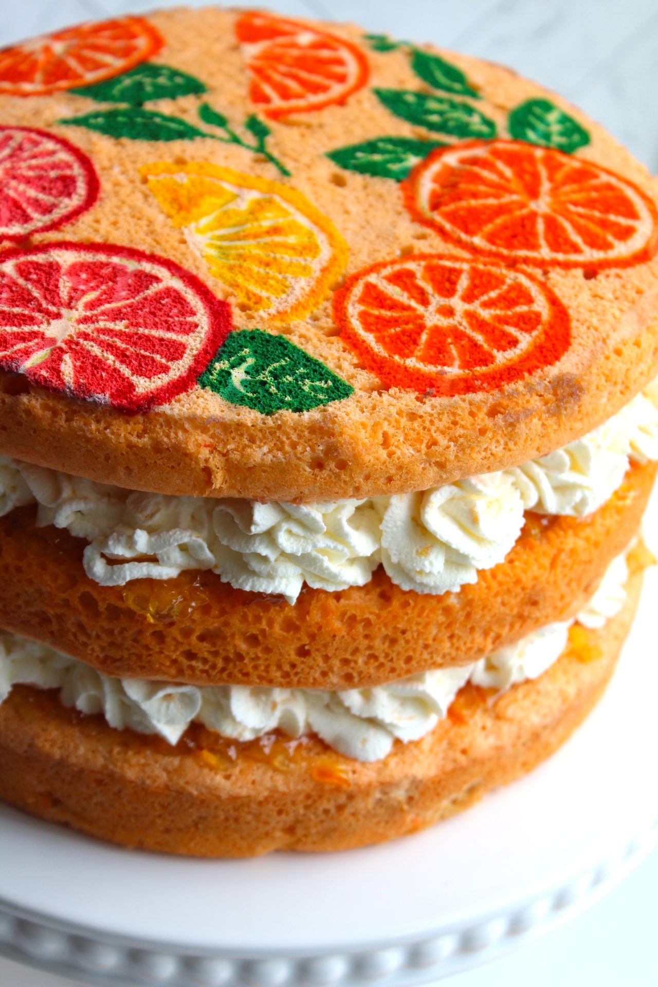 A three-layer sponge cake with cream and fruit filling between the layers. The top layer and sides of the cake are decorated with vibrant, colorful prints of various citrus fruits, including oranges, lemons, and grapefruits. The cake is displayed on a white cake stand.