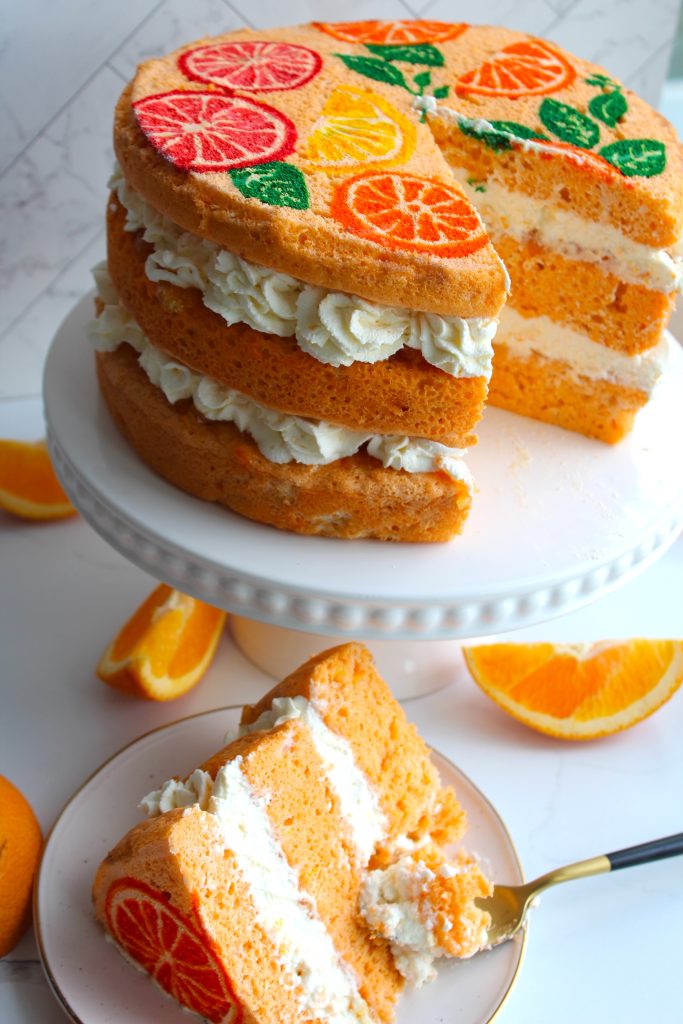 A three-layer orange cake topped with colorful citrus fruit decorations and filled with white cream sits on a white cake stand. A slice is placed on a plate in front, with a fork beside it, showing the creamy layers amidst the vibrant orange sponge.