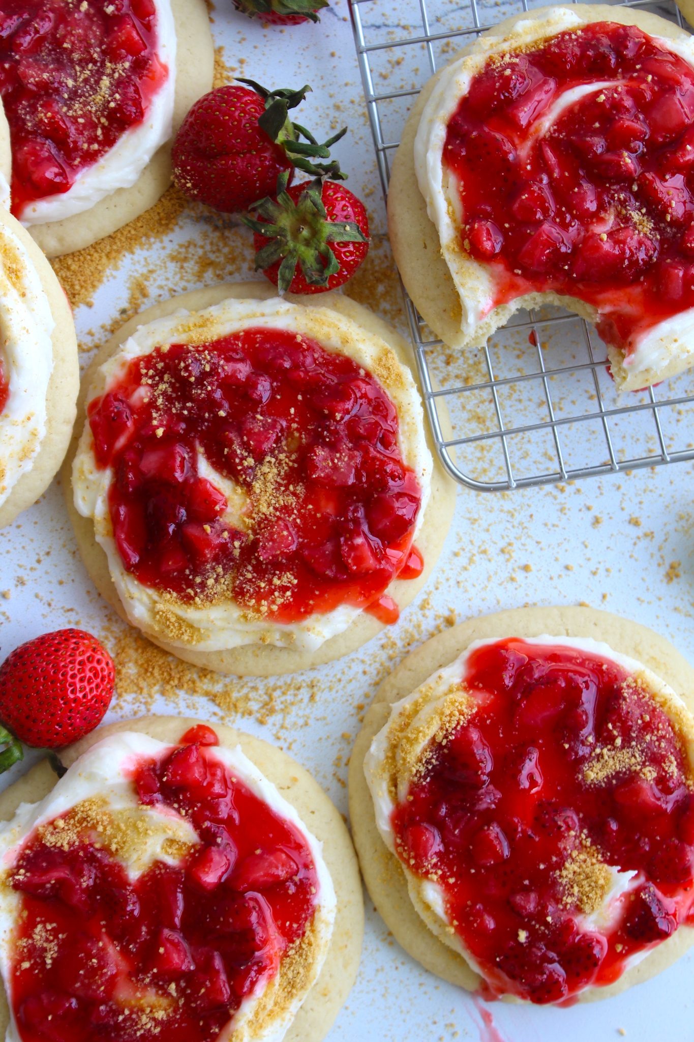Close-up of several sugar cookies topped with cream cheese frosting and strawberry sauce, with some fresh strawberries around. One cookie has a bite taken out of it and rests on a wire cooling rack, while the others are on a white surface sprinkled with crumbs.