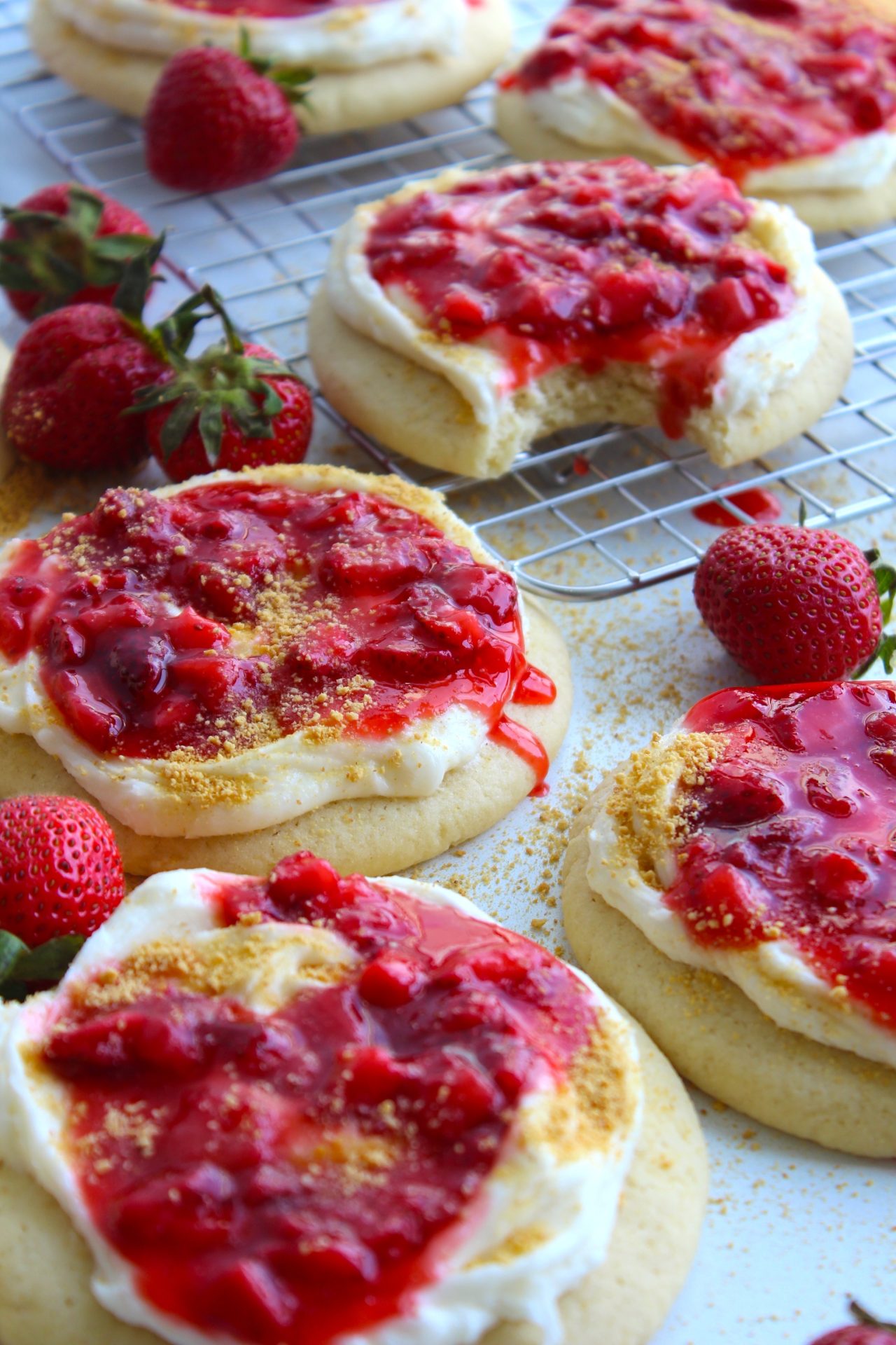 Several strawberry-topped cookies with a creamy white frosting are displayed on a cooling rack and a white surface. Fresh strawberries are scattered around the cookies. The cookies are garnished with a crumbly topping, and one cookie has a bite taken out of it.