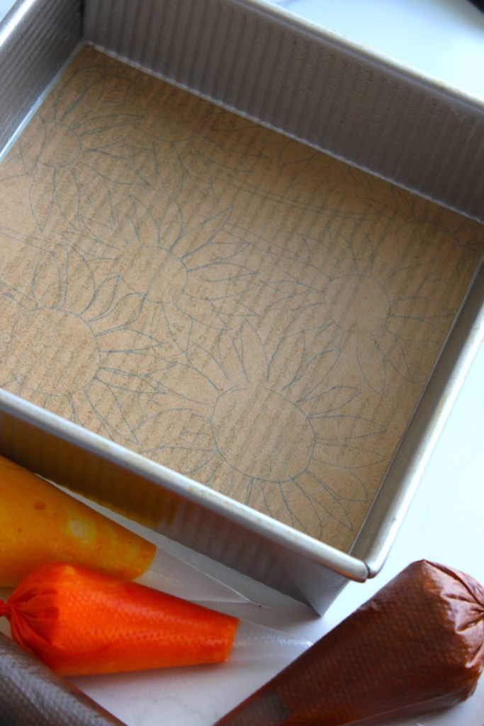 A square baking pan lined with parchment paper, which has sunflower patterns drawn on it. Three piping bags filled with colorful icings, orange, yellow, and brown, are placed nearby on a white surface.