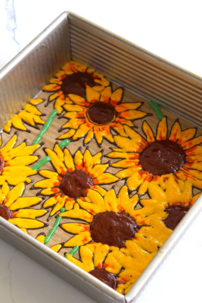 A square baking pan contains a batter that has been decorated with a vibrant sunflower design. The flowers have bright yellow petals with brown centers, and some green stems and leaves can be seen. The pan is set on a white surface.