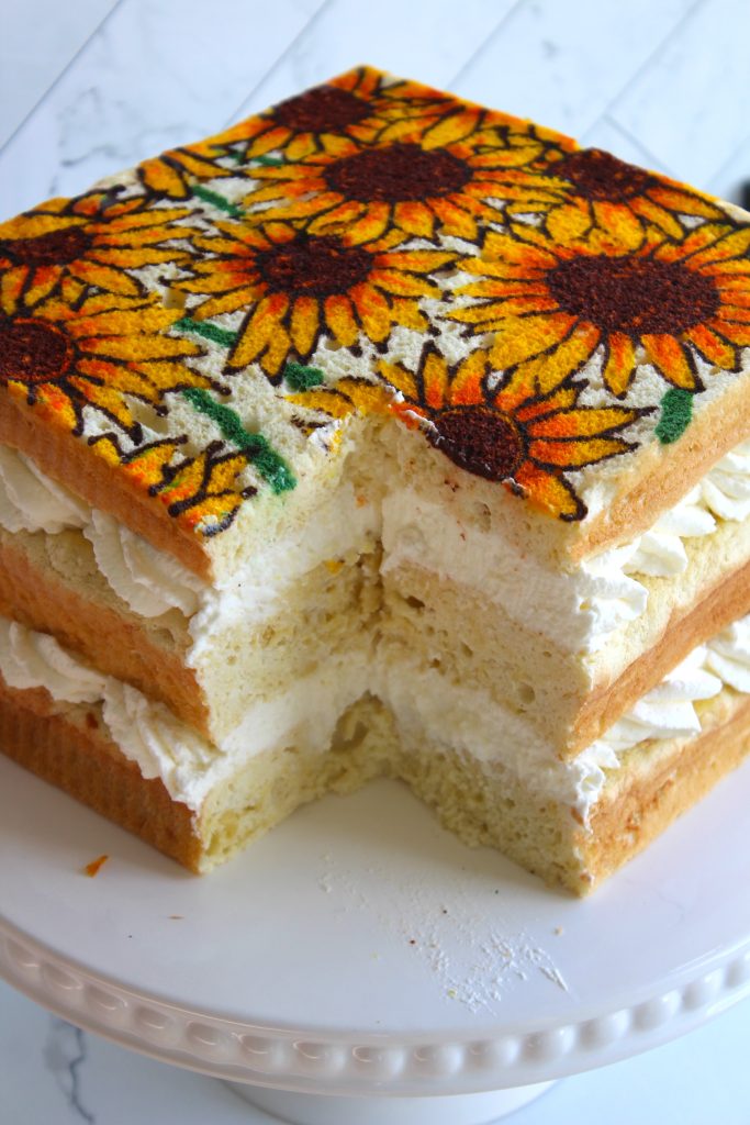 A square sponge cake with three layers of cream filling is topped with a vibrant design of sunflowers. The cake sits on a white cake stand, and a slice has been cut out, revealing the fluffy layers and cream inside. The background is a white marble surface.