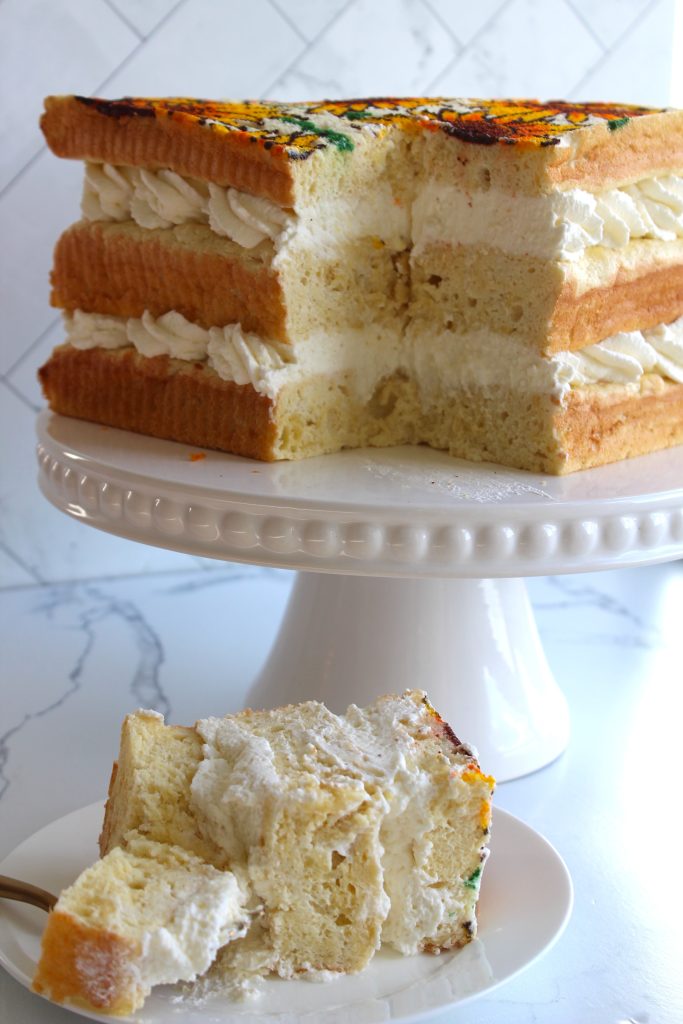 A three-layer cake with thick layers of white frosting sits on a white cake stand. A large slice has been cut and is displayed on a small white plate in front of the stand. The cake features decorative yellow and orange flowers on the top layer.