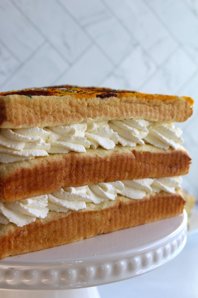 A close-up shot of a layered dessert featuring three layers of sponge cake with swirls of whipped cream in between. The dessert is presented on a white cake stand against a blurred, white-tiled background.