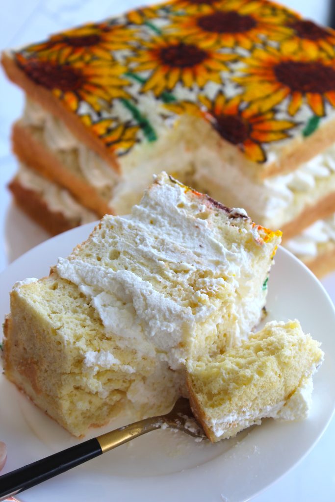 A fork is cutting into a slice of layered cake covered in whipped cream on a white plate. The cake slice is from a larger cake in the background, which has a decorative sunflower design on top.