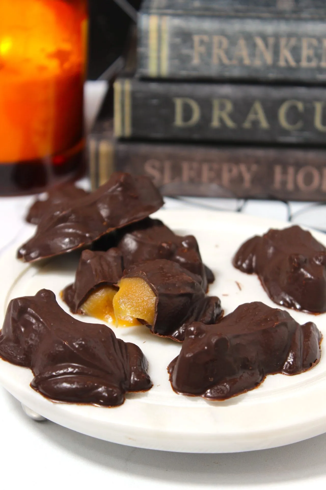 A white plate holds several chocolate frogs, some of which are broken open to reveal a caramel filling. In the background, spooky-themed books with titles "Frankenstein," "Dracula," and "Sleepy Hollow" are stacked next to an orange candle.