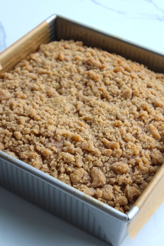A square metal baking pan filled with a freshly baked crumb cake, featuring a golden-brown, crumbly topping. The cake remains in the pan, resting on a parchment paper lining, with a light background barely visible around the edges.