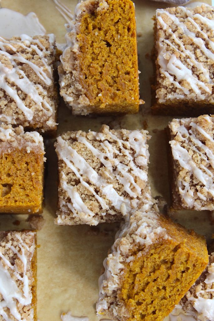 Close-up of nine square slices of orange pumpkin crumb cake drizzled with white icing, arranged neatly on parchment paper. The cakes have a crumbly top and moist interior, with visible layers and texture.