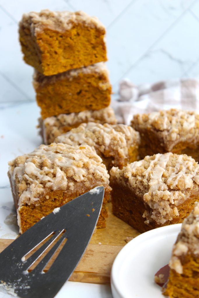 A close-up of several slices of moist pumpkin coffee cake with crumb topping and glaze. One slice is being lifted with a spatula, while others are stacked in the background. A checkered cloth wraps around some of the slices in the background.
