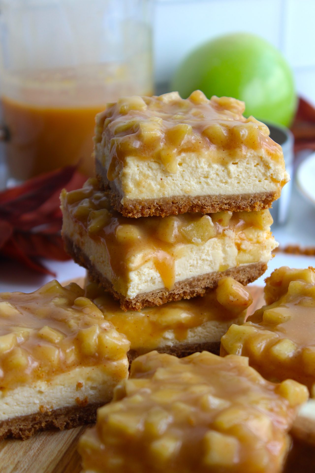A stack of caramel apple cheesecake bars with a buttery crust, creamy cheesecake layer, and a rich caramel apple topping. The bars are placed on a wooden surface with a blurred green apple and a jar of caramel in the background.