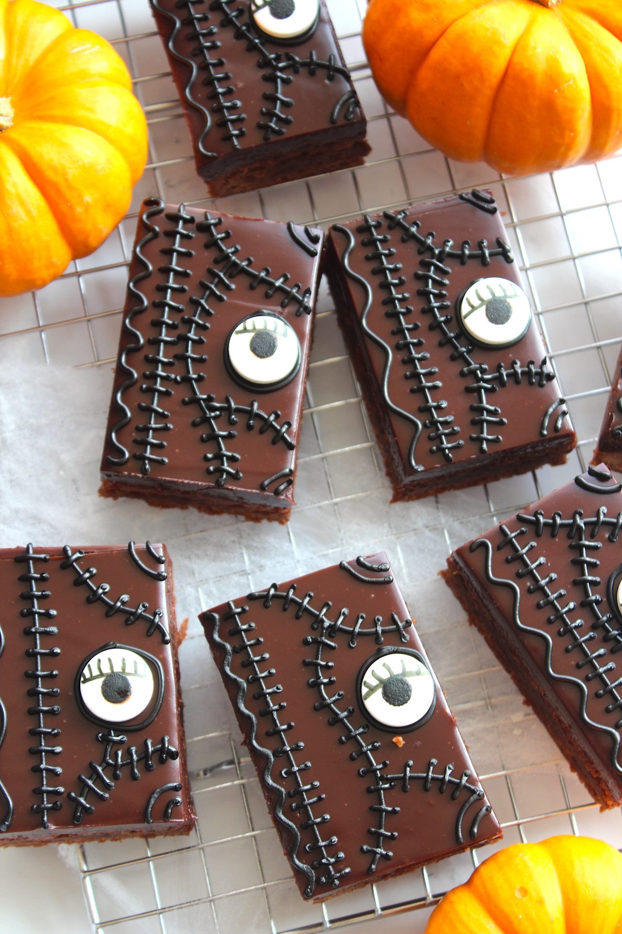 Chocolate brownies decorated with creepy black frosting stitches and candy eyeballs, resembling a stitched monster face, are arranged on a wire rack. Small pumpkins are placed around the brownies, adding to the Halloween theme.