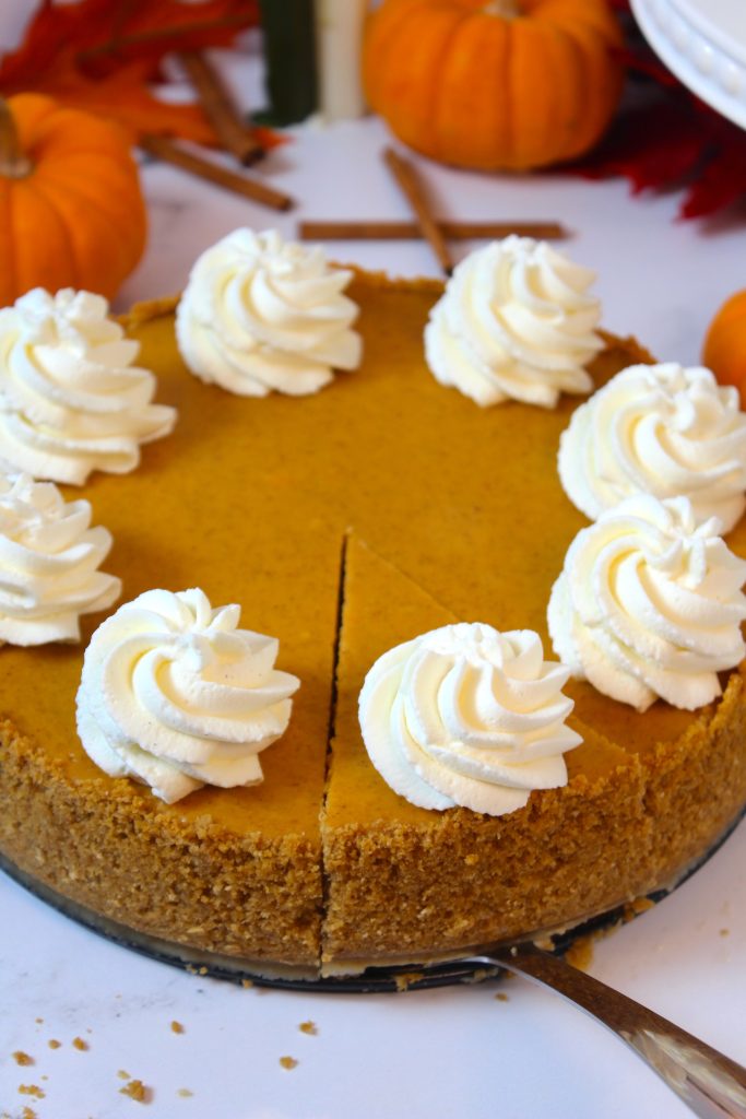 A pumpkin cheesecake with a graham cracker crust is topped with swirls of whipped cream. The cheesecake is partially sliced, and several small pumpkins and cinnamon sticks are in the background, creating an autumnal scene.