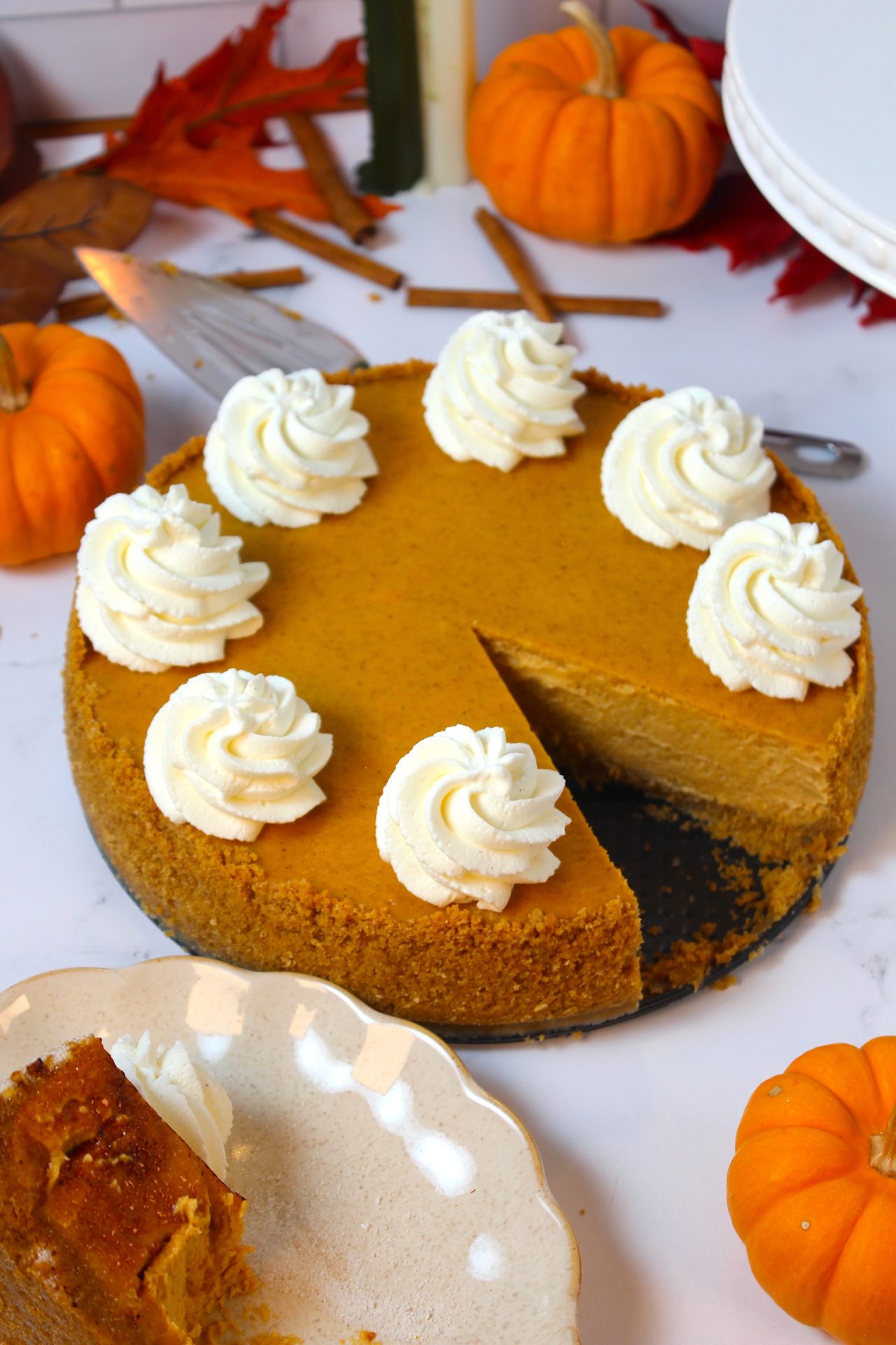 A pumpkin cheesecake topped with eight swirls of whipped cream sits on a white surface surrounded by small pumpkins, cinnamon sticks, and autumn leaves. A slice has been cut out and is placed on a white plate in the foreground.