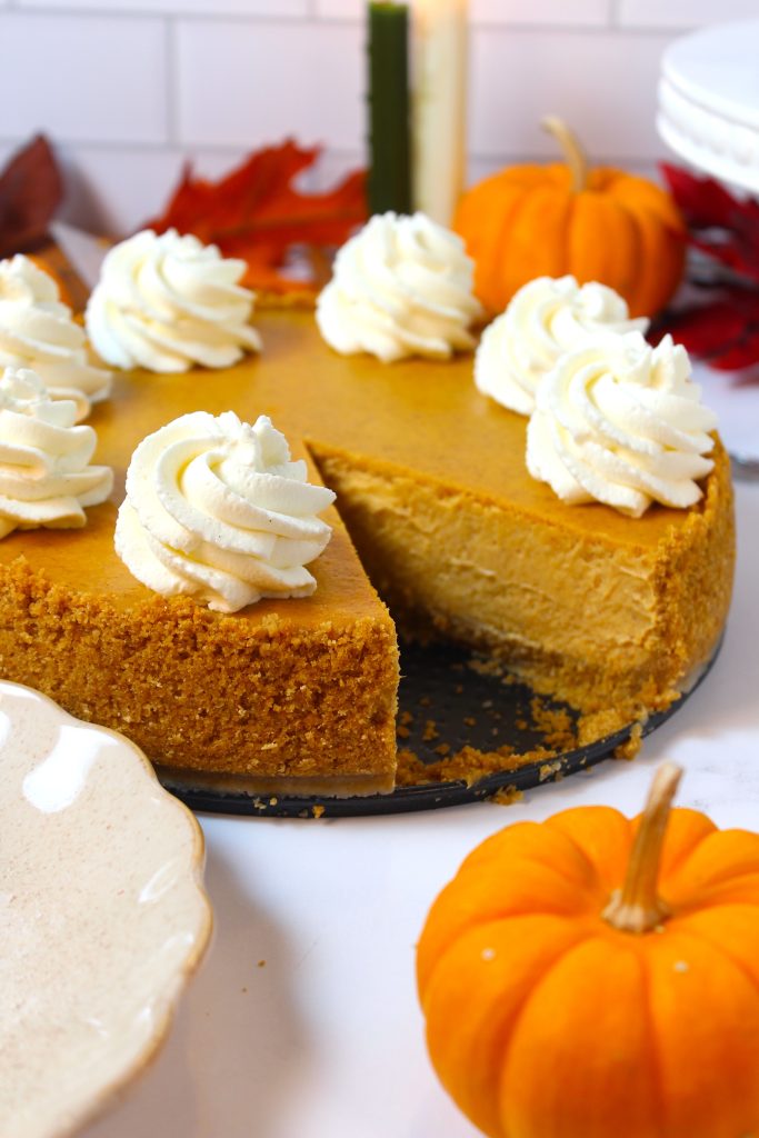 A pumpkin cheesecake topped with swirls of whipped cream is presented on a table. One slice is removed, revealing the creamy interior. The table is adorned with small pumpkins, autumn leaves, and a white plate, suggesting a fall or Thanksgiving theme.