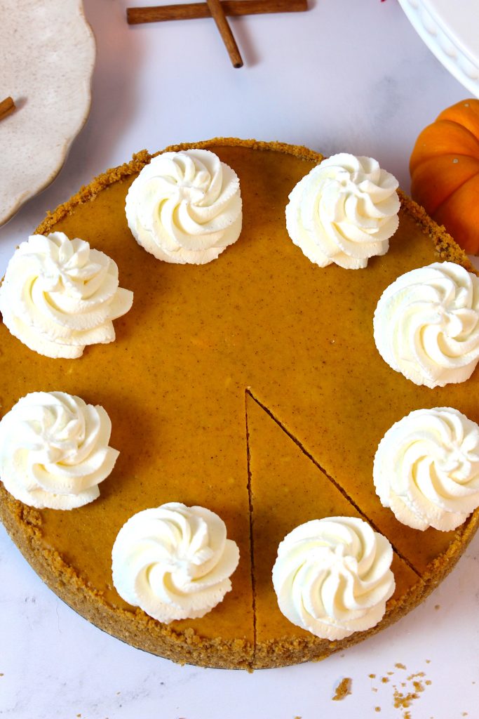 A pumpkin pie topped with eight swirls of whipped cream is shown on a white surface. One slice has been cut from the pie. The pie has a light brown crust and smooth orange filling. Nearby, there are cinnamon sticks and a small orange pumpkin.
