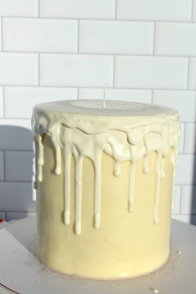 A tall, round white cake is covered with smooth frosting and topped with glossy white icing that drips down the sides. The cake is set against a white tiled background.