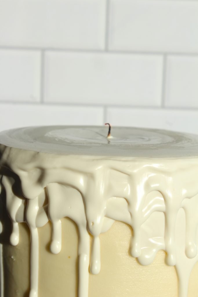 A close-up of a white frosted cake with smooth icing drips down the sides. The top has a single exposed candle wick. The background features a tiled wall, giving a clean and simple look.