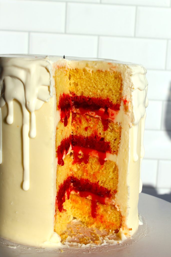 A four-layer cake with white icing and red filling between the layers. The top and sides have white icing drips. The cake is on a white surface with a faintly visible tiled wall in the background.
