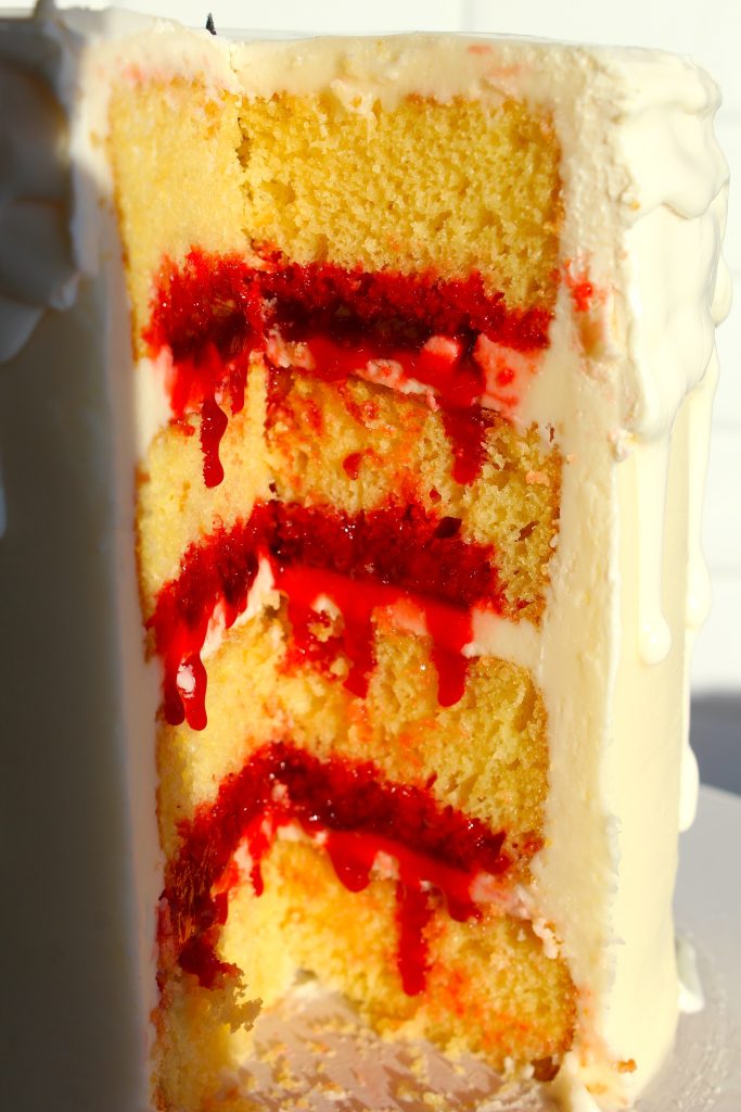 A sliced cake with several yellow layers interspersed with red filling and white icing. Sunlight highlights the textures, making the red filling appear vibrant against the soft, creamy icing.