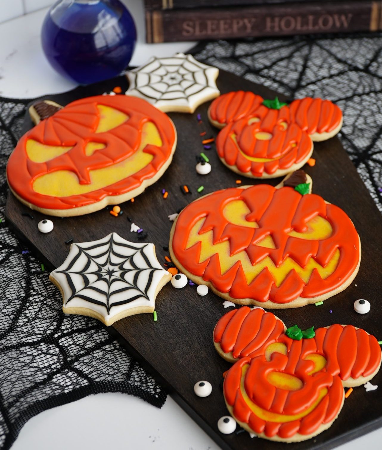 A tray with Halloween-themed cookies, including jack-o’-lanterns and Mickey Mouse-shaped pumpkins iced in orange, and spiderweb cookies. The tray is on a black lace cloth, with books and a purple potion bottle in the background.
