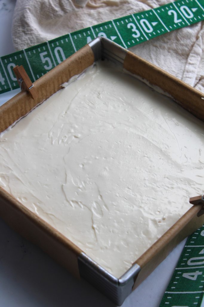 A square baking pan filled with smooth, creamy batter. The pan is lined with parchment paper and secured with small clips at the corners. A green measuring tape and a beige cloth are nearby on a white surface.