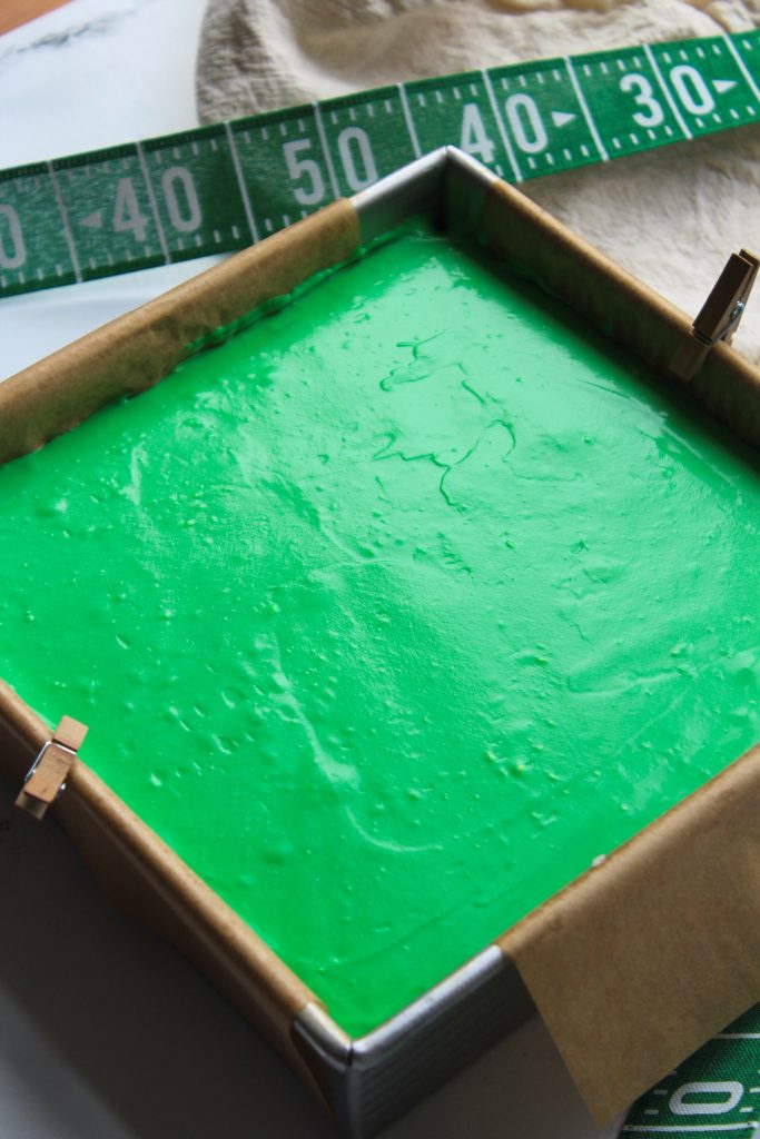 A square baking pan filled with green batter, placed on parchment paper with a measuring tape marked with yard lines in the background.