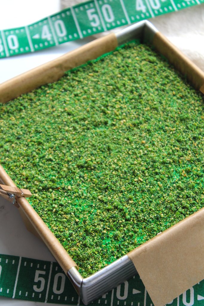 A rectangular baking pan filled with green, grass-like textured cake decorated to resemble a football field. A measuring tape with yard markers is partially visible around the pan's edge.