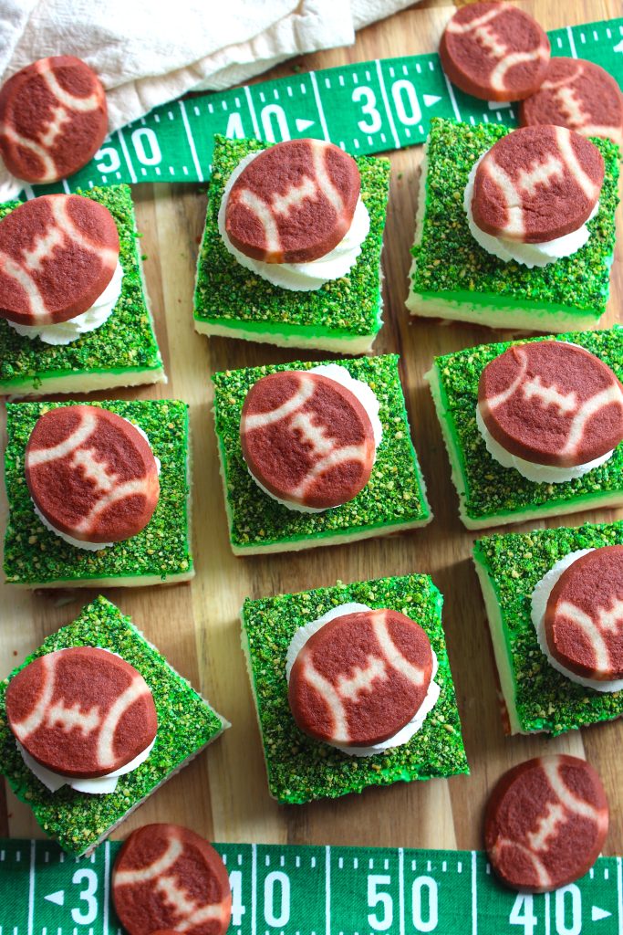 Top view of green frosted square treats topped with white frosting and football-shaped cookies, placed on a football field-themed board.