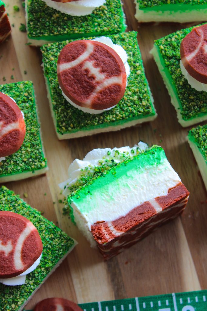 Various green and white layered dessert bars topped with football-shaped cookies and green sprinkles on a wooden surface. One dessert is sliced to reveal its inner layers, and a football field design is partially visible underneath.