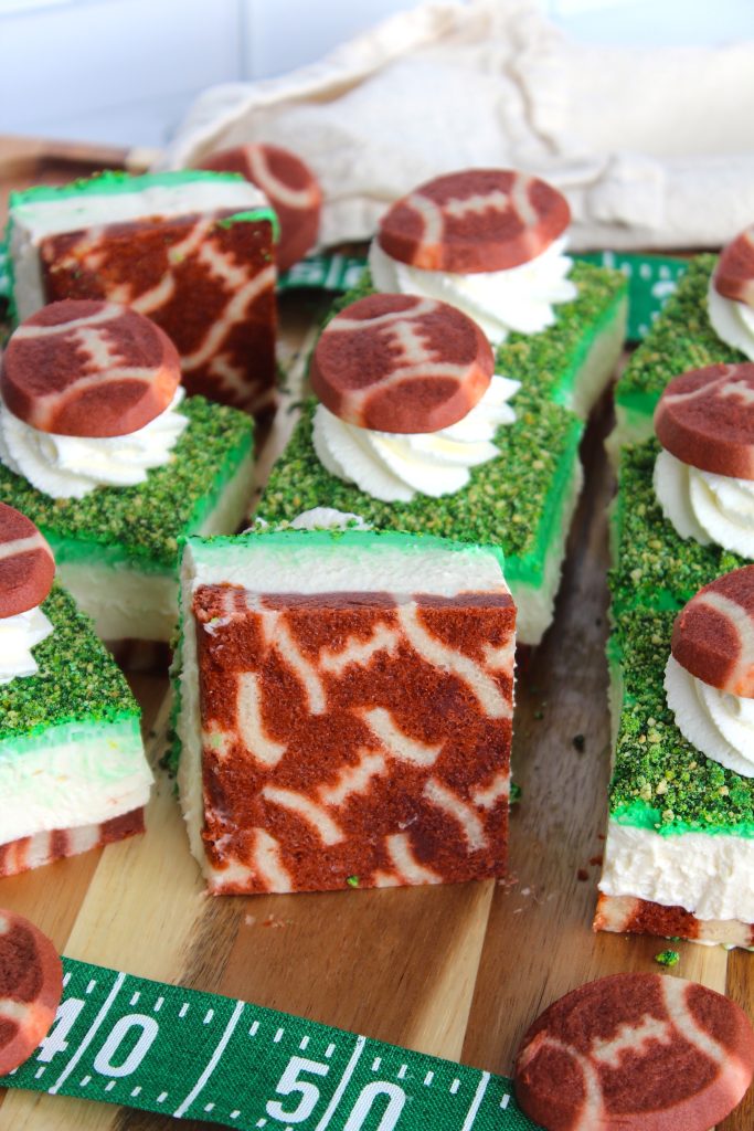 Sliced brownies decorated with green icing and grass-like sprinkles, topped with whipped cream and football-shaped cookies. A ruler-like strip in shades of green lies nearby, resembling a sports field.