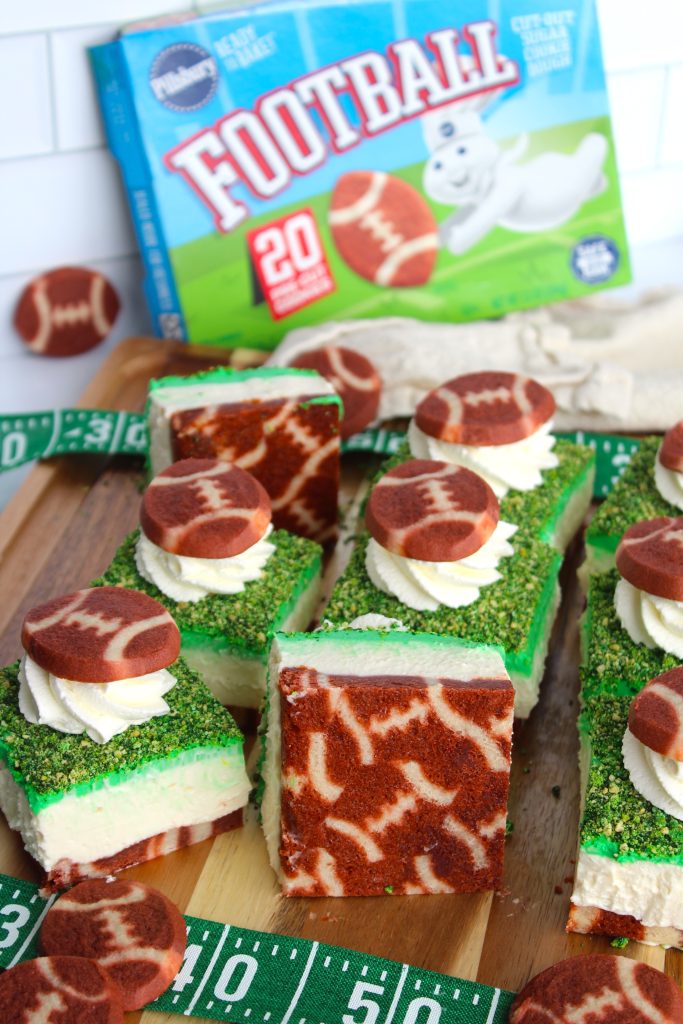Dessert squares topped with football-shaped cookies and whipped cream are presented on a wooden board. They have green icing and a marbled cake layer. A box with baking mix labeled "Football" is in the background. Football-themed decor surrounds the setup.
