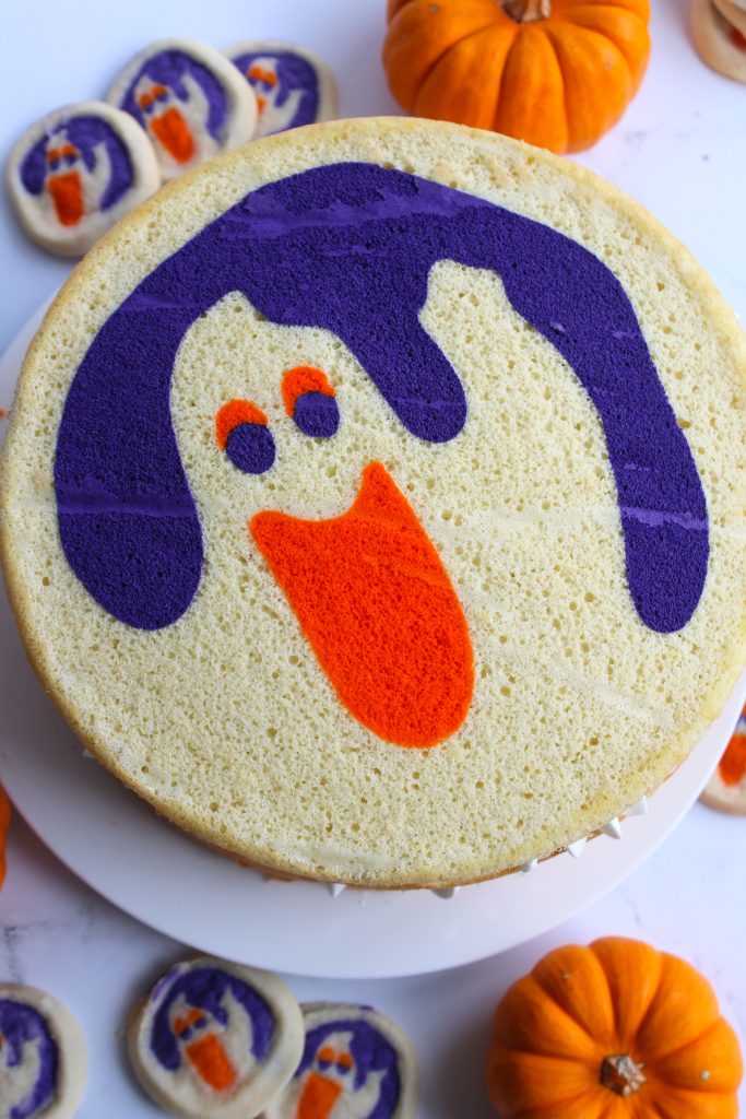 A round Halloween cake features a cheerful ghost with purple hair and an orange mouth. Surrounding the cake are small pumpkin decorations and ghost-themed cookies.