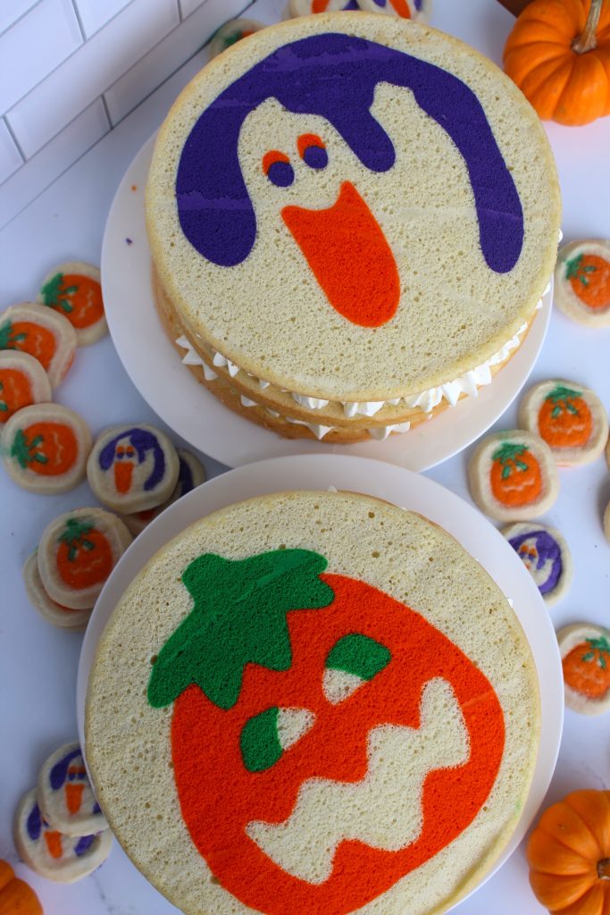 Two Halloween-themed cakes with playful ghost and pumpkin designs. Surrounding the cakes are assorted decorated cookies featuring similar spooky motifs. The scene is set on a white surface with small pumpkins in the background.