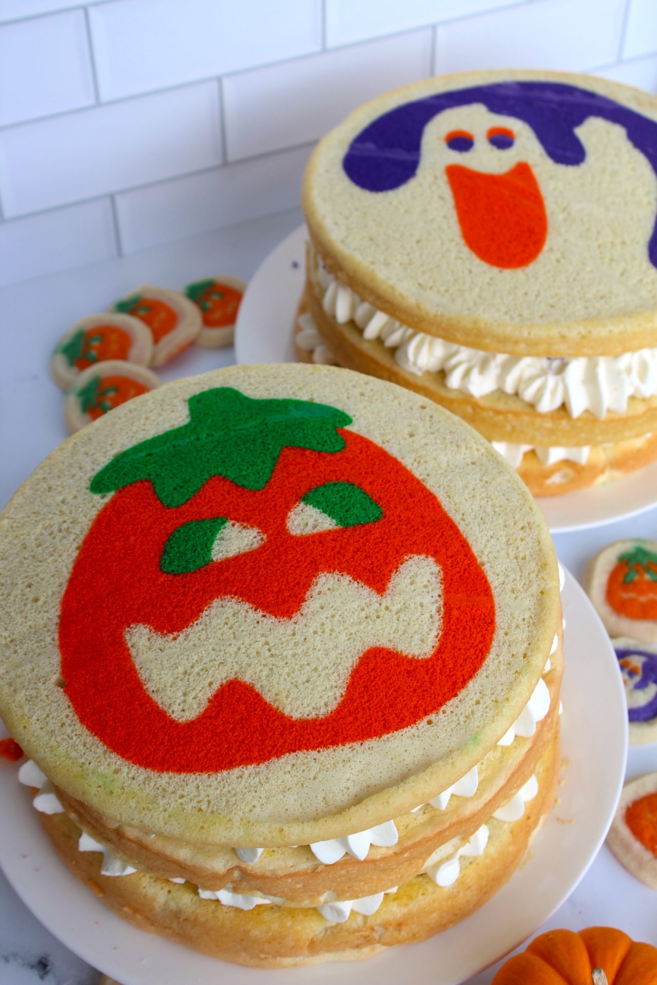 Two decorated sponge cakes with whipped cream filling are displayed. One cake features a red jack-o'-lantern design on top, while the other shows a purple ghost. Small pumpkin-themed cookies are on the side.